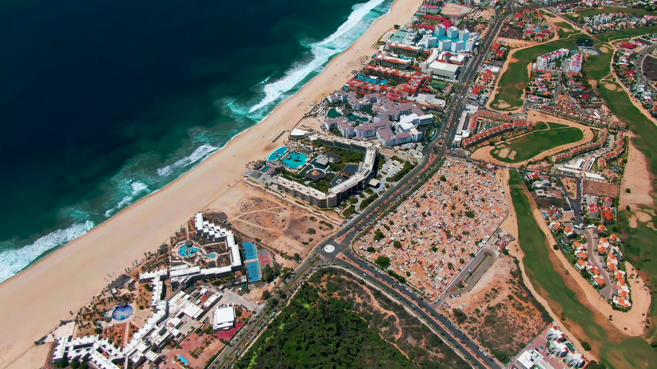 Opening aerial frame over San Jose Del Cabo, Baja California Sur, with dark ocean and surf at left, sandy beach, coastal road, resort buildings, golf course greens, and no visible people.