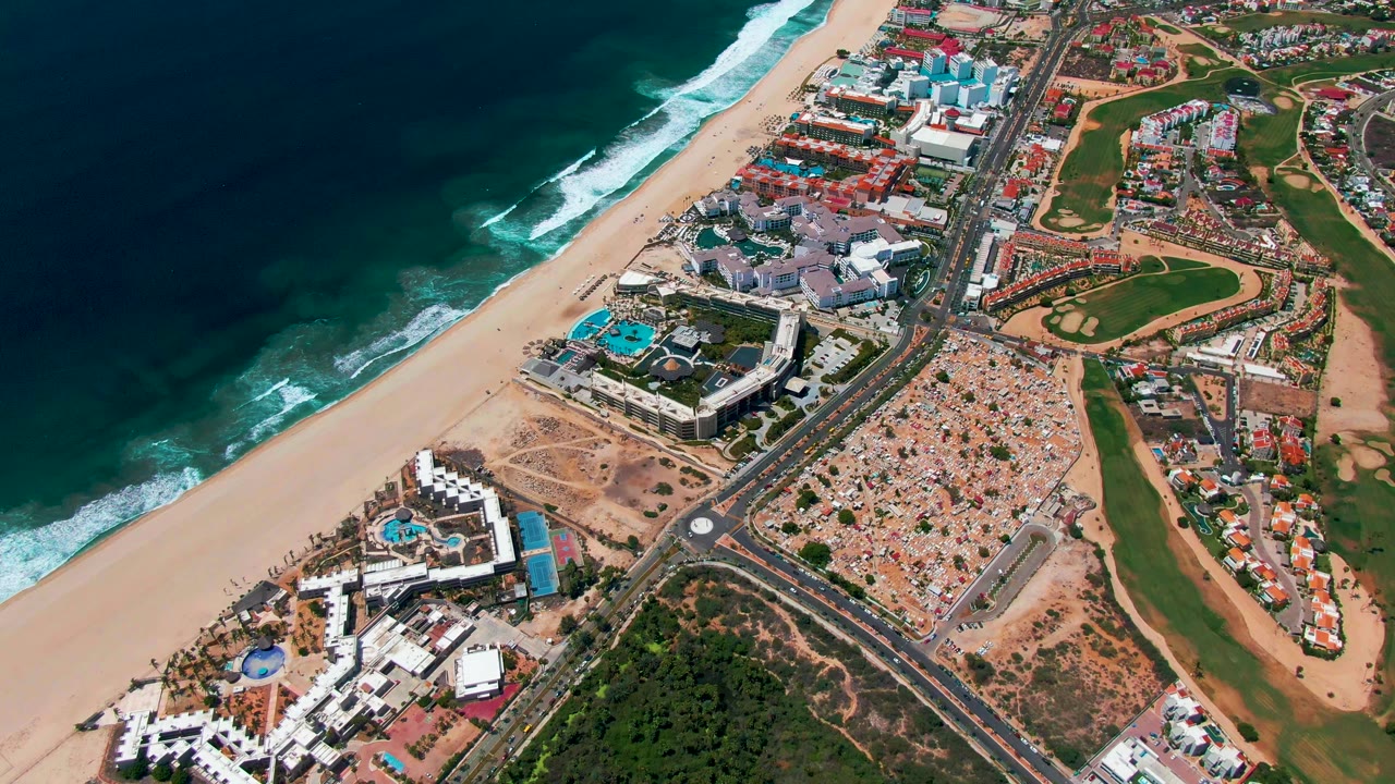 San Jose Del Cabo aerial view one second in, with waves breaking beside the beach, a road and roundabout through the center, clustered buildings, golf course fairways, and no visible people.