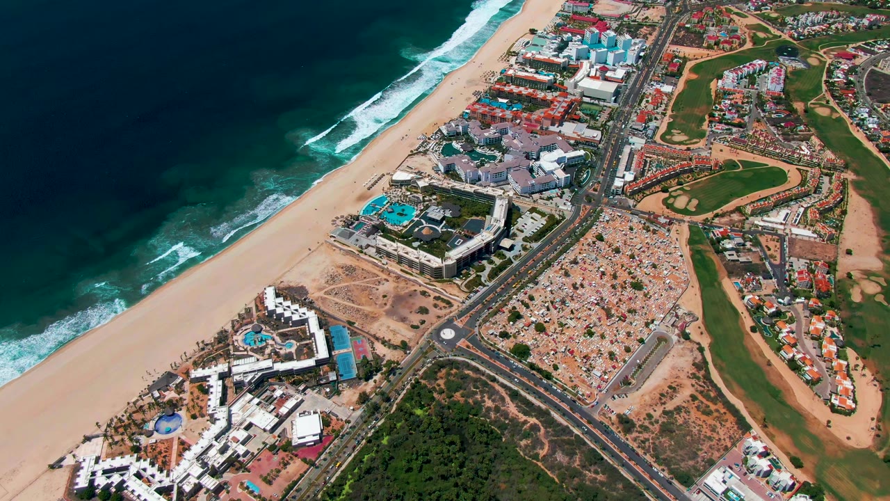 Two seconds into the San Jose Del Cabo aerial, the shoreline runs diagonally with white surf, beach sand, a coastal road carrying small vehicles, resort buildings, golf course areas, and no visible people.
