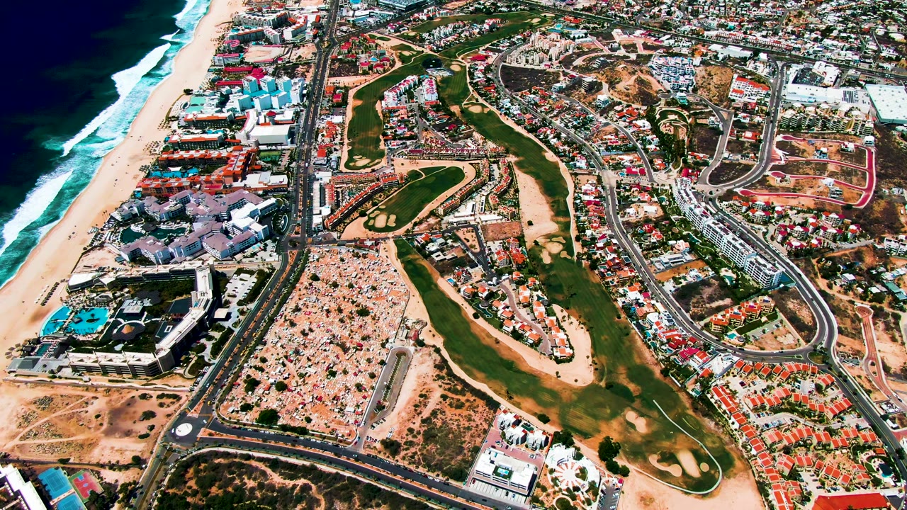 Opening aerial frame over San Jose Del Cabo, Baja California Sur, with ocean waves on the left, a sandy beach, central golf fairways, roads, resort buildings, residential blocks, and no visible people.