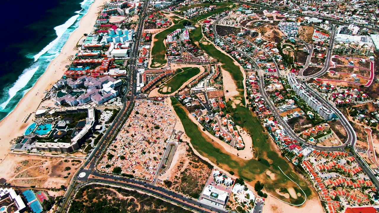 Oblique drone view of surf, sand, fairways, roadways, and dense coastal development.