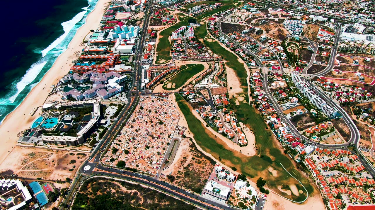 Mid-clip aerial over San Jose Del Cabo with dark blue ocean, bright breaking waves, a pale beach strip, golf greens, tan undeveloped patches, resort complexes, housing clusters, and no visible people.