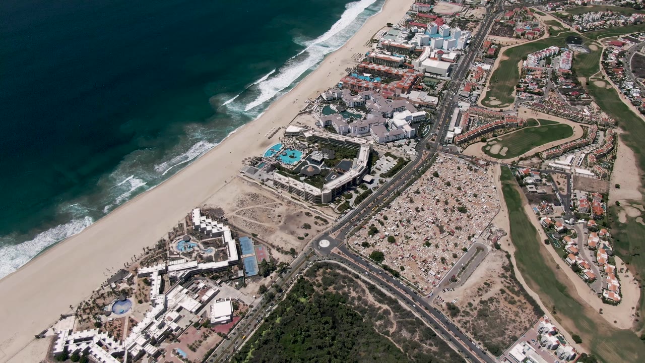 Second frame of the San Jose Del Cabo aerial view, with waves shifted along the beach edge and cars visible on the divided coastal road, with no visible people.