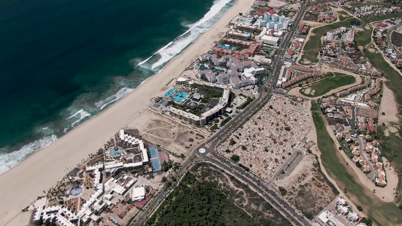 Aerial San Jose Del Cabo frame with dark ocean water, white surf lines, a broad sandy beach, active road lanes, clustered buildings, and golf course greens, with no visible people.