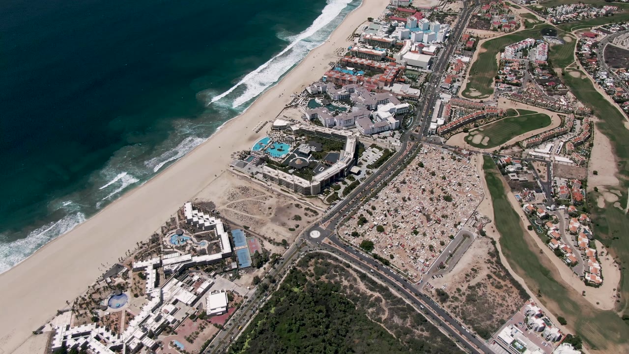 Aerial coastline with ocean surf, beach, road intersection, cars, buildings, and golf course.