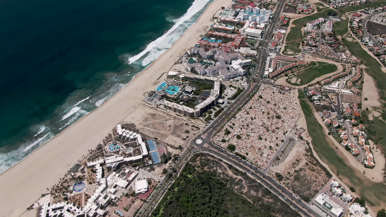 Aerial view over San Jose Del Cabo with dark blue ocean, white surf, a sandy beach, beachfront buildings, a road, golf course greens, and no visible people.