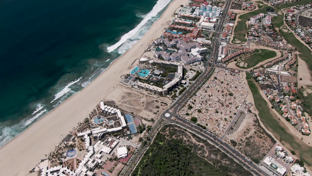 Drone view over San Jose Del Cabo with ocean waves on the left, a wide beach, clustered beachfront buildings, a curved road, golf course areas, and no visible people.