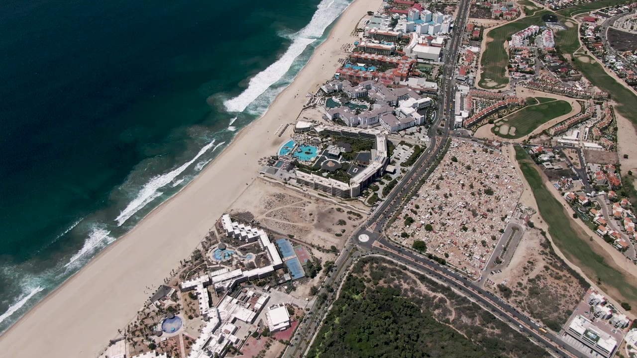 Aerial view of ocean surf, sandy beach, coastal road, buildings, and green areas.