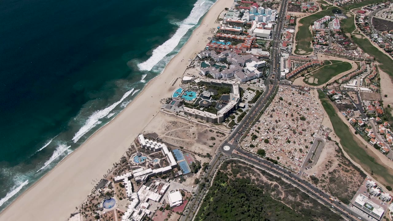 Aerial frame over San Jose del Cabo, Mexico, with white wave lines edging the beach, clustered buildings near the sand, a road corridor, green areas, and no visible people.