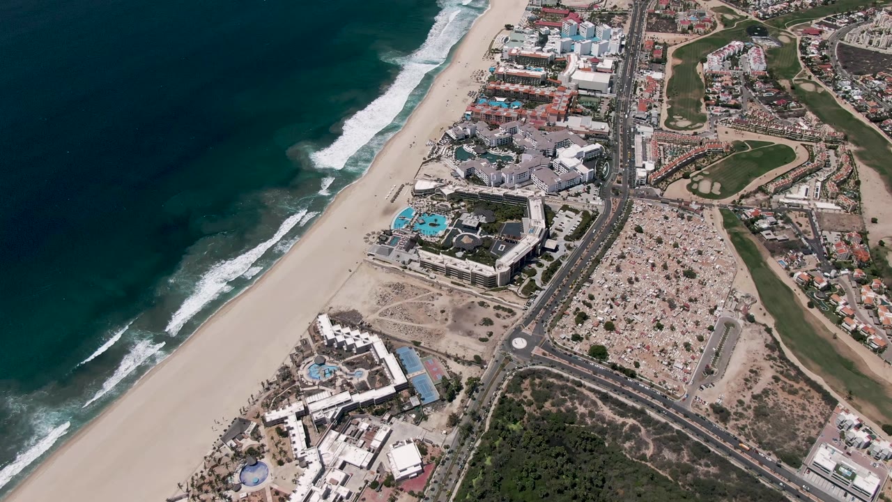 San Jose del Cabo aerial frame showing foamy surf on the left, empty-looking sand, a built-up beachfront area, road traffic, green landscaping, and no visible people.