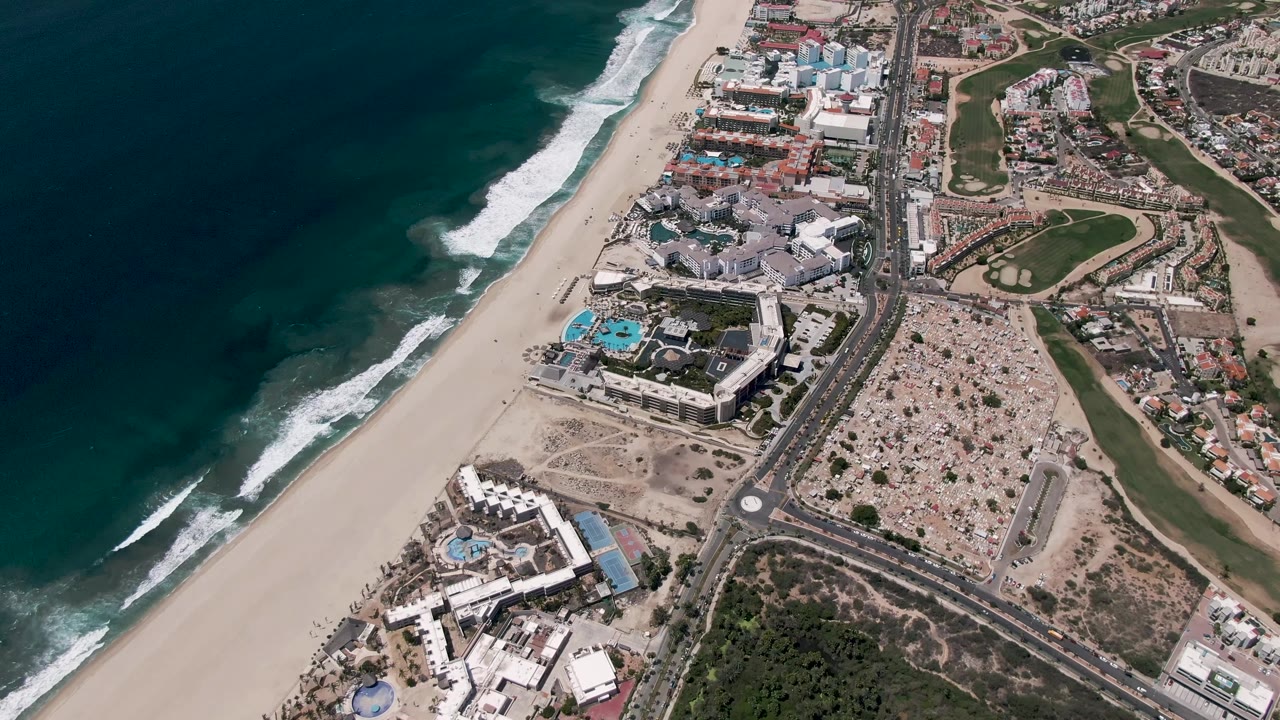 Mid-clip overhead view of San Jose del Cabo, Mexico, with the ocean and surf band to the left, sandy shore below, road junctions, buildings, greenery, and no visible people.