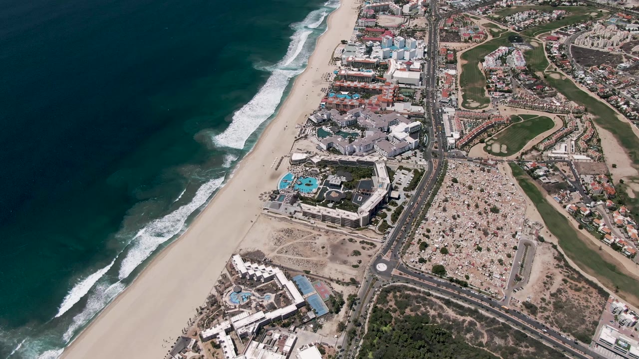 Aerial view over San Jose Del Cabo with dark blue ocean at left, white surf on a wide sandy beach, coastal buildings and a road inland, and no visible people.