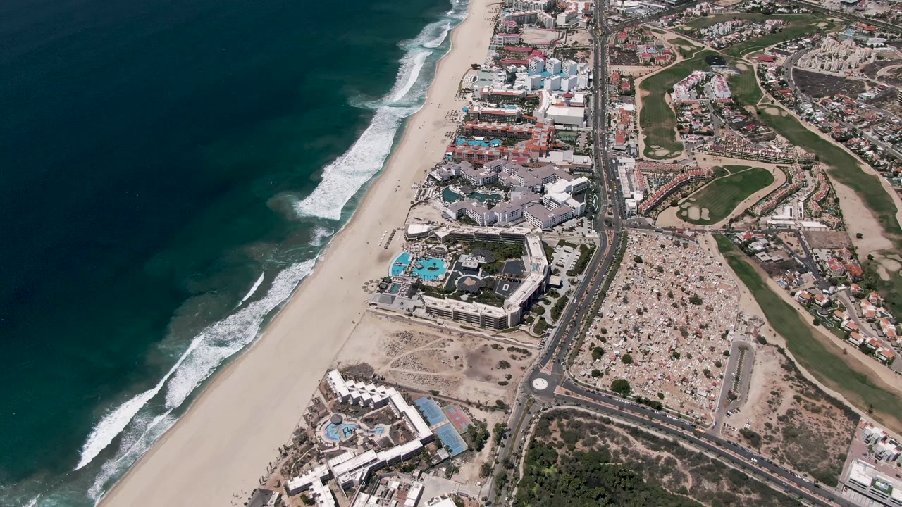 Aerial San Jose Del Cabo coastline frame with the surf line slightly shifted along the beach, turquoise pools among coastal buildings, a road, golf greens, and no visible people.