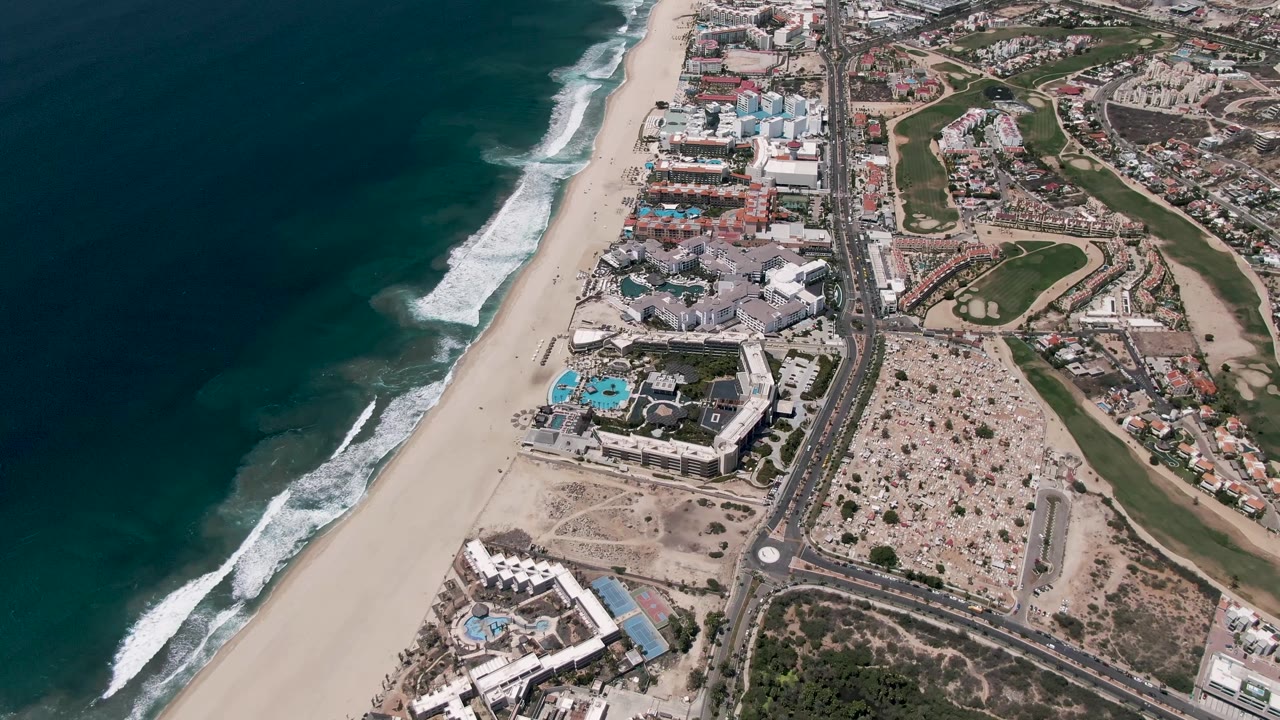 High aerial view of San Jose Del Cabo where white waves reach farther up the sandy shore beside a coastal road, clustered buildings, golf greens, and no visible people.