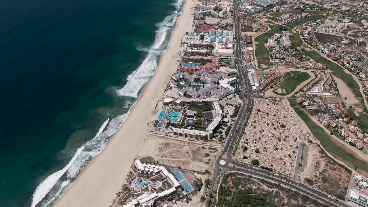 Oblique aerial frame over San Jose Del Cabo with a broader run of ocean visible at left, surf breaking diagonally onto sand, coastal buildings and golf greens inland, and no visible people.