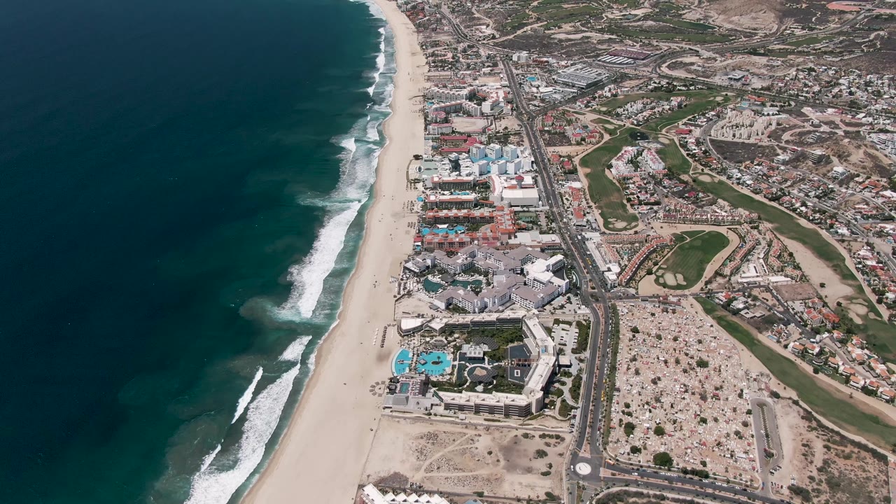 Aerial frame from San Jose del Cabo, Baja California Sur, showing blue-green ocean, white surf, a broad sandy beach, coastal buildings, roads, parking areas, and no visible people.