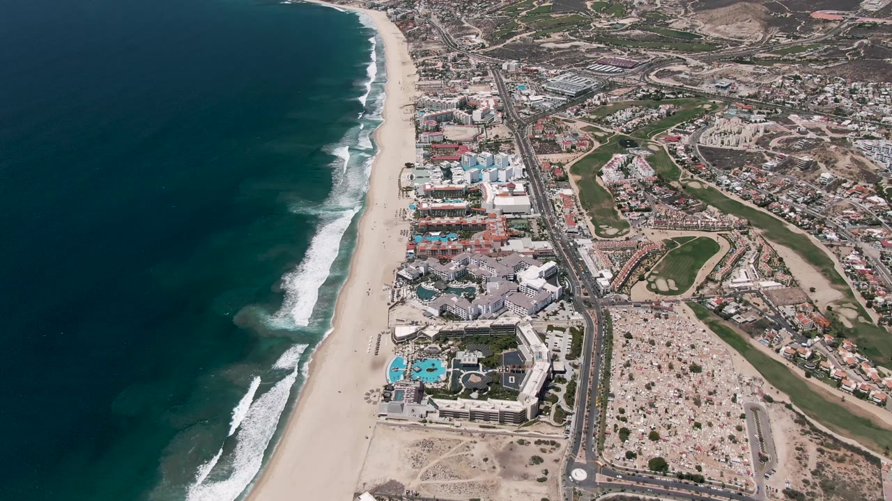 A later aerial frame of San Jose del Cabo shows the whitecap pattern shifted along the shore, with ocean on the left, sand in the center, buildings, roads, parking, and no visible people.