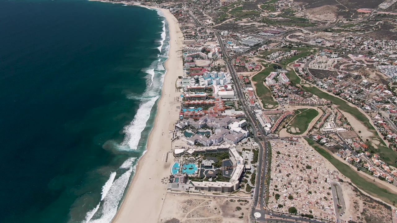 Aerial view over San Jose del Cabo with surf bands closer to the beach, a pale sand strip, beachfront structures, a main road, parking areas, inland development, and no visible people.