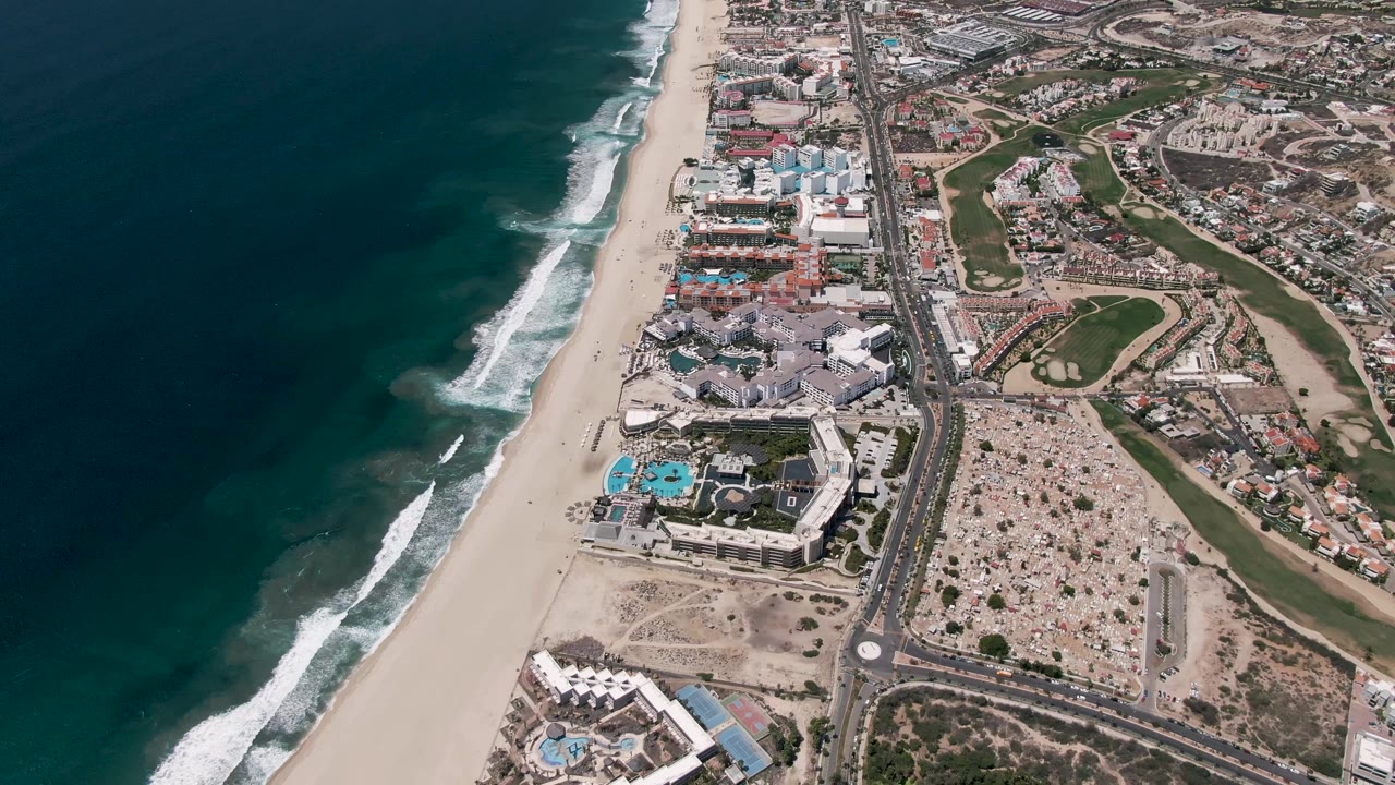 High aerial view of San Jose del Cabo coastline at the start of the clip, with dark blue ocean at left, white surf on the sand, a parallel coastal highway, resort pools, buildings, green fairway areas, and no visible people.