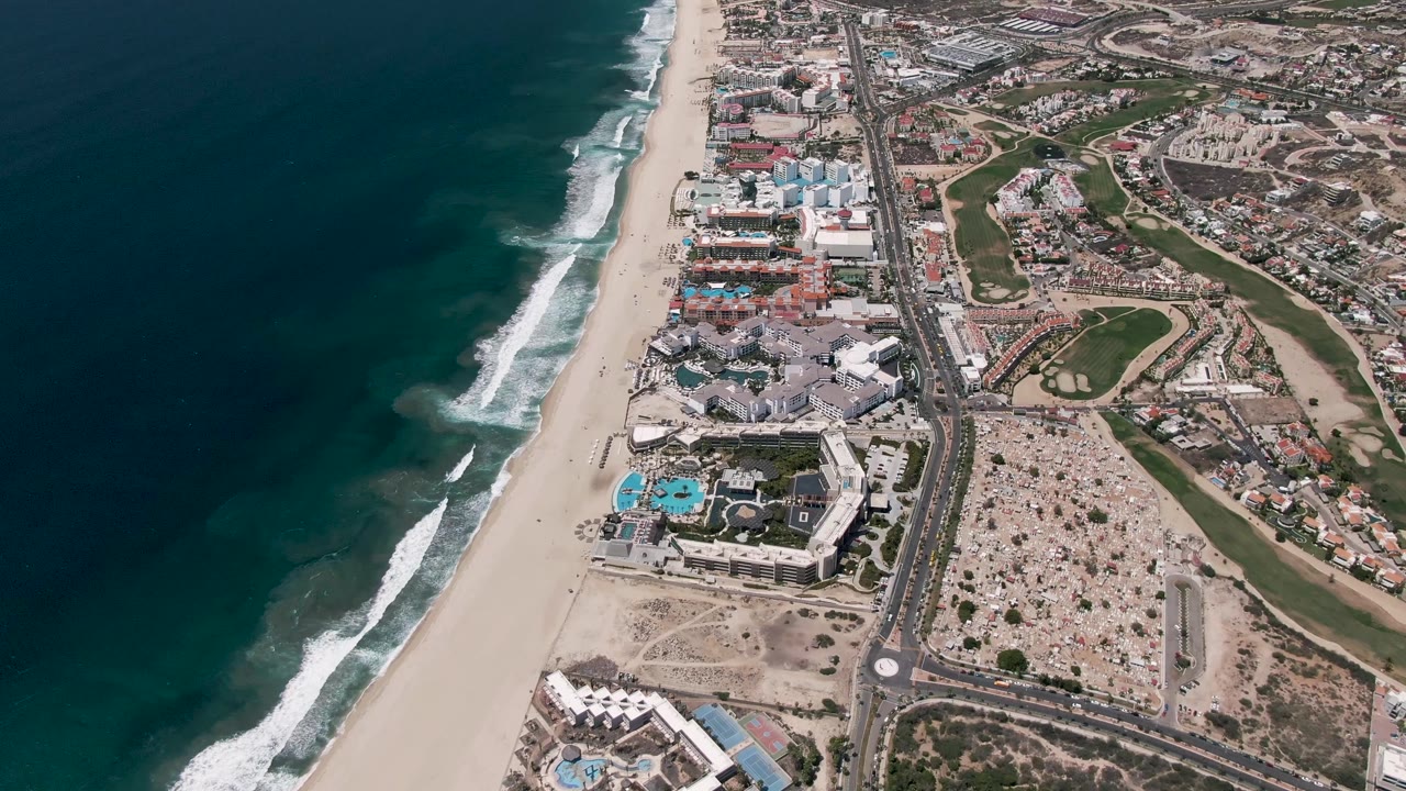 San Jose del Cabo aerial frame one second in, showing waves advancing along the beach, a coastal highway just inland, clusters of resort buildings and blue pools, landscaped green areas, and no visible people.