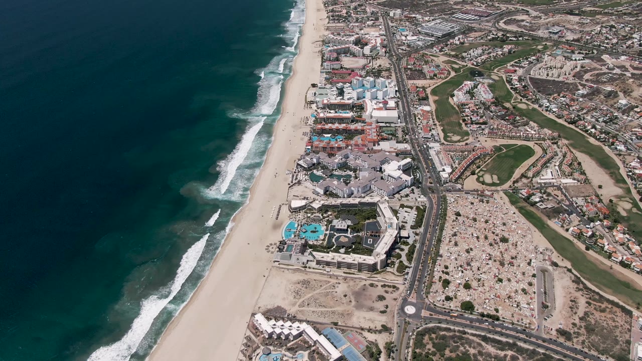 San Jose del Cabo coastline from above at 0:02, with the ocean mass on the left, white wave bands nearing shore, a pale sand beach, a road corridor, resort pools, built-up lots, and no visible people.