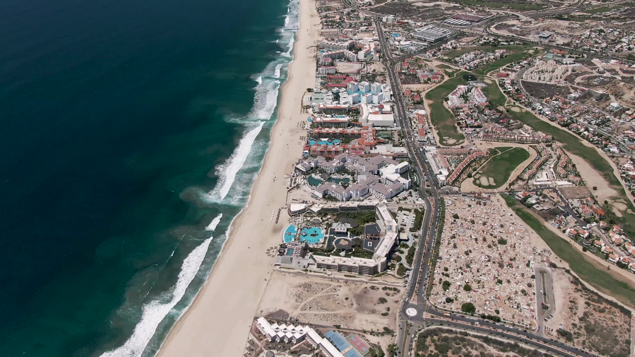 Mid-clip aerial view over San Jose del Cabo, where foamy waves line the beach, the highway follows the shoreline, resort pools sit near the lower beach edge, inland buildings fill the right side, and no visible people appear.