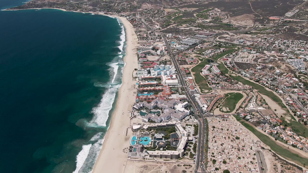 Aerial frame over San Jose Del Cabo with dark blue ocean on the left, white surf along the sand, resort buildings and pools near the beach, and no visible people.