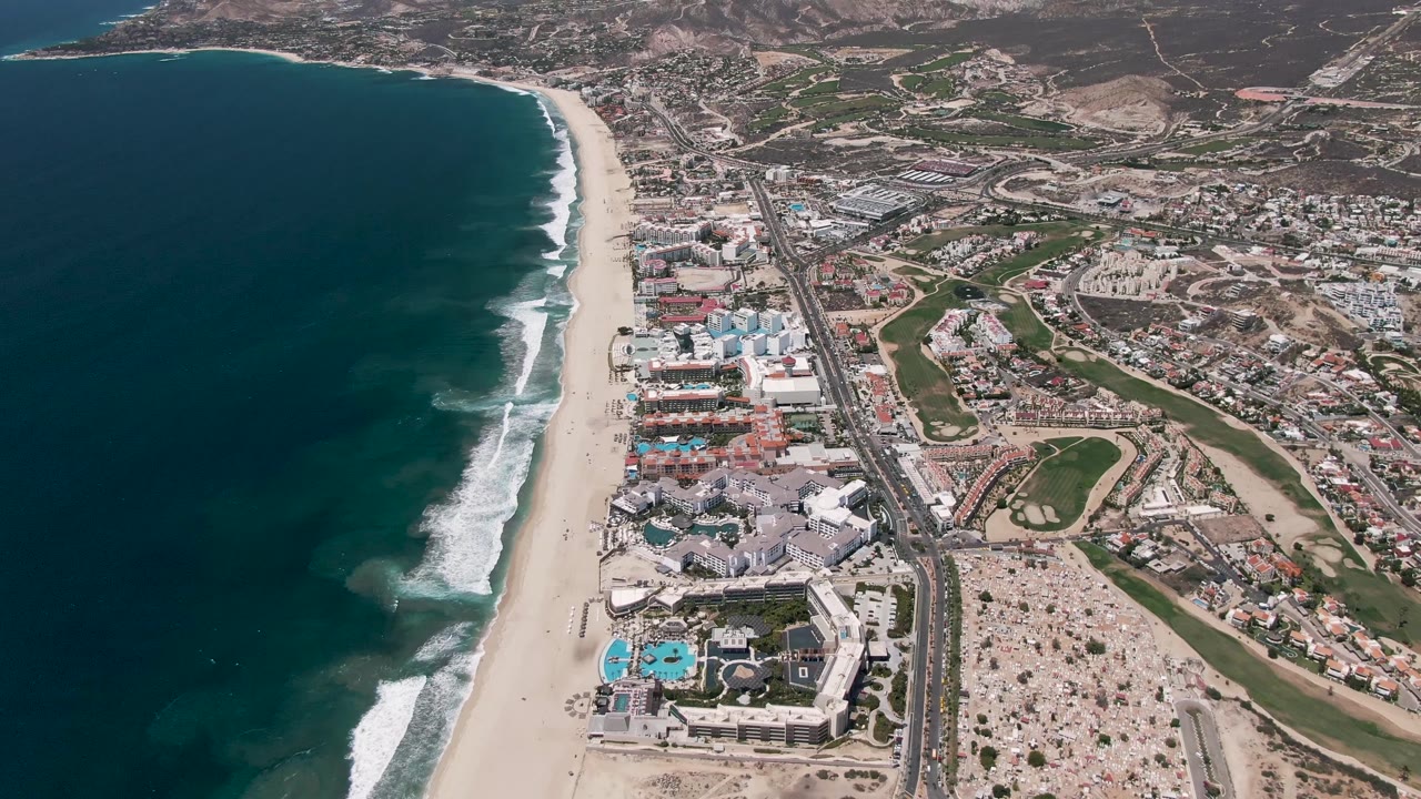 San Jose Del Cabo aerial view with the surf line slightly advanced along the beach, a road running near the shore, resort-style buildings, pools, and no visible people.