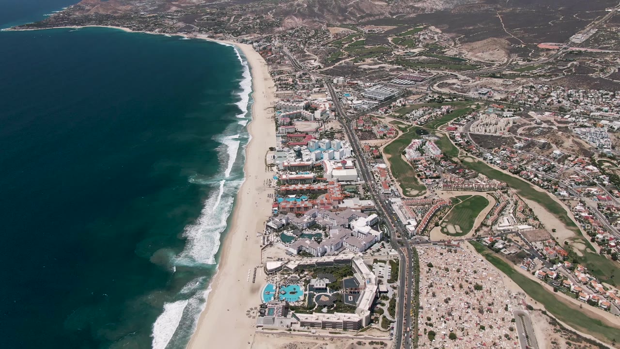 A high aerial view in San Jose Del Cabo shows white foam bands rolling toward the sandy beach, with coastal buildings, bright pools, a straight road, and no visible people.