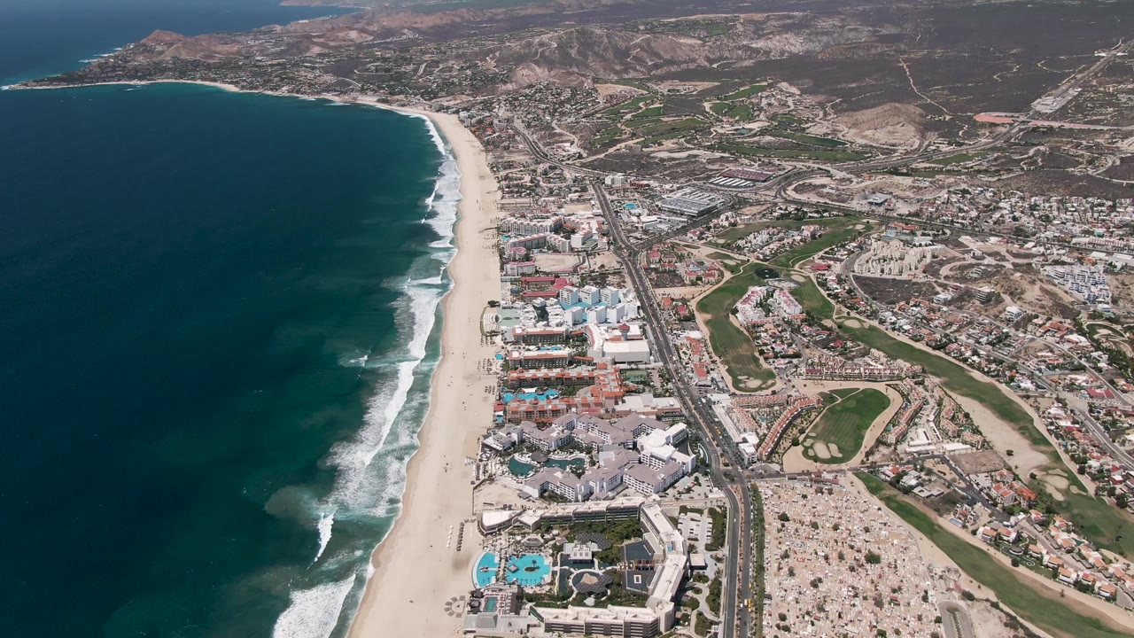 High aerial frame over San Jose Del Cabo, Mexico, with dark ocean at left, a pale sandy beach, a parallel highway, coastal buildings, green spaces, dry hills, and no visible people.
