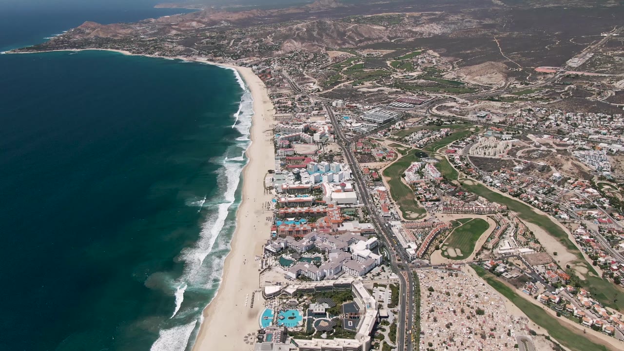 Aerial coastline with surf, highway traffic, buildings, and dry hills.