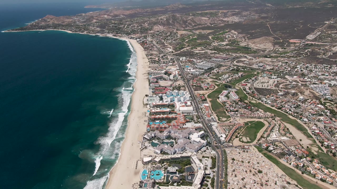 Wide aerial frame over San Jose Del Cabo with teal water and white wave bands on the left, beach and highway in the middle, dense coastal buildings to the right, dry terrain beyond, and no visible people.