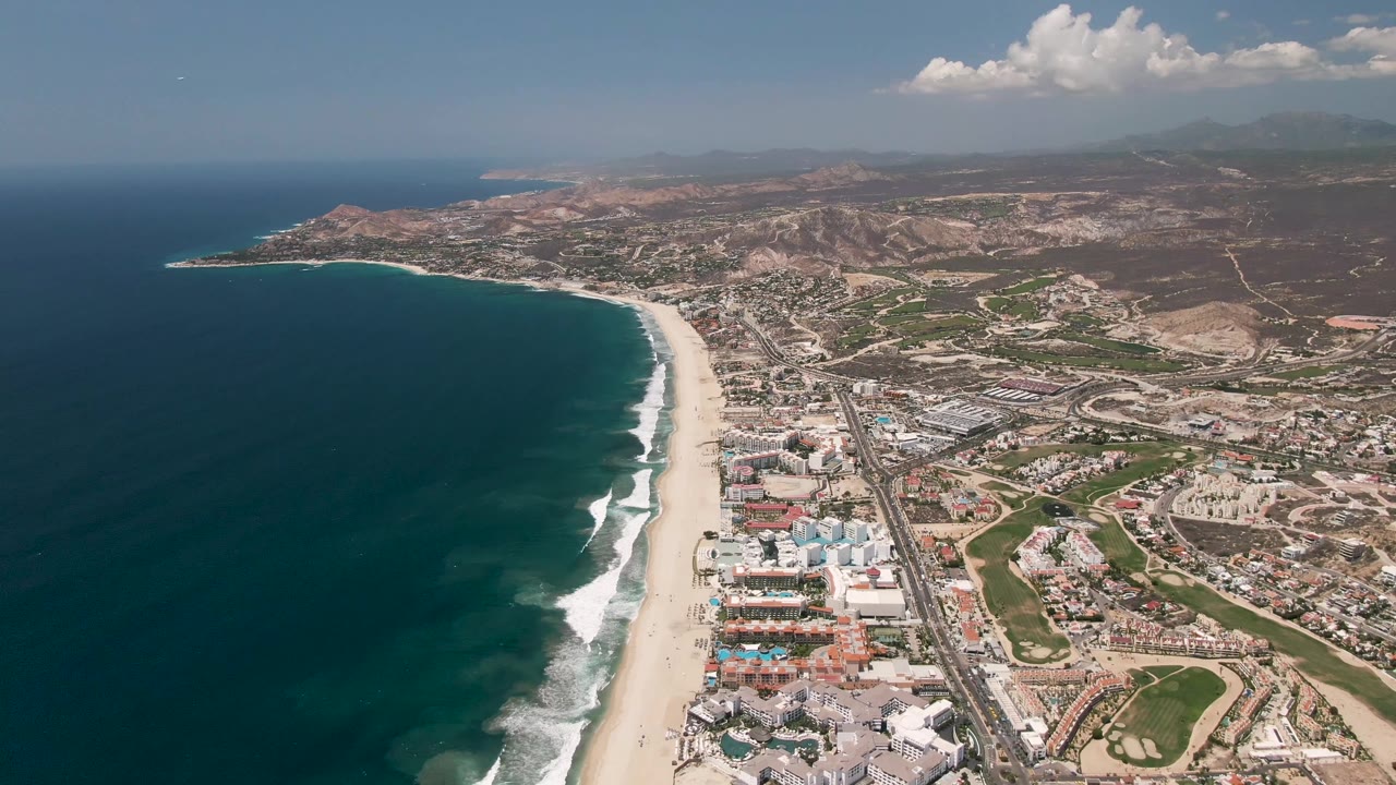 Wide aerial view of San Jose del Cabo coastline with deep blue ocean at left, a sandy beach and surf line, a coastal highway, developed land, hills, clouds, and no visible people.