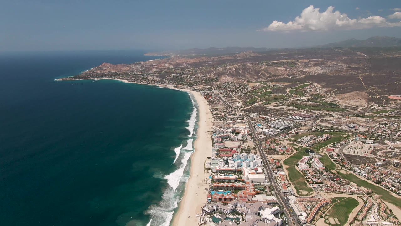 Aerial frame over San Jose del Cabo with the ocean filling the left side, white surf along the beach, a parallel road corridor, clustered buildings, dry hills, clouds, and no visible people.