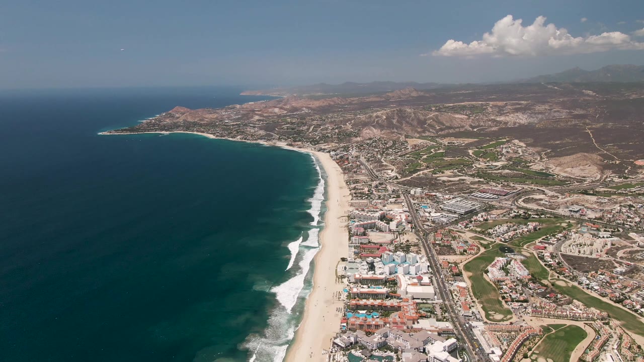 High aerial view of San Jose del Cabo coastline where the curved beach, rolling surf, coastal highway, resort-style buildings, inland roads, hills, and no visible people remain in frame.