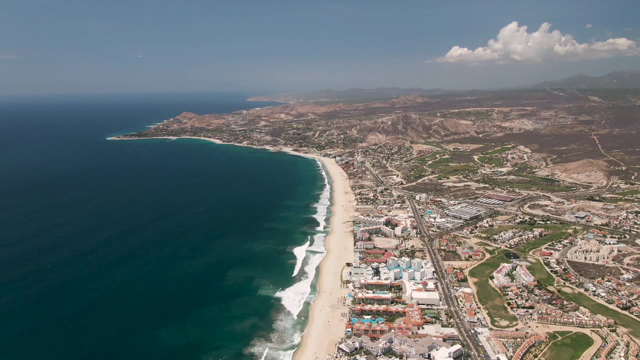 Wide drone view of ocean, beach, highway, coastal buildings, and mountains