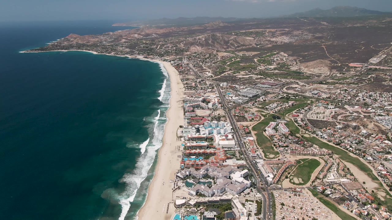 Opening aerial frame over San Jose del Cabo with dark ocean on the left, a long surf-lined beach, urban buildings beside a highway, inland hills, and no visible people.