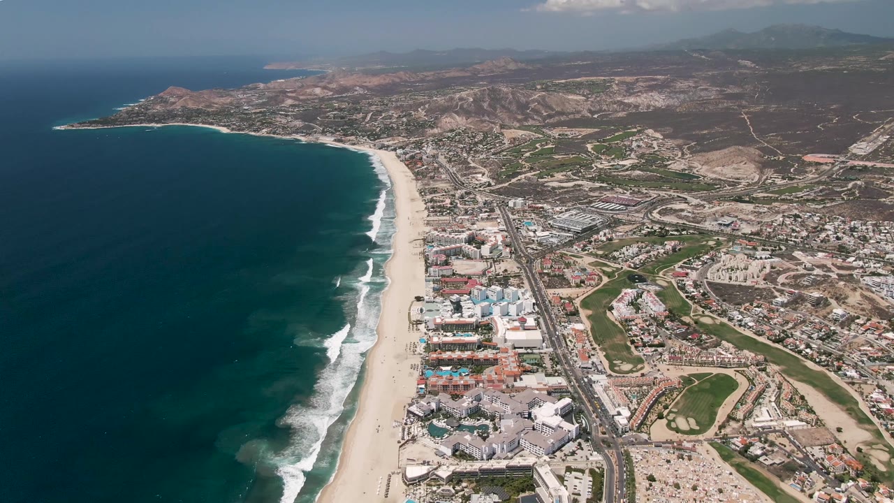 San Jose del Cabo aerial frame showing the curving beach and white surf closer to center, with beachfront buildings, a straight highway corridor, dry hills, and no visible people.