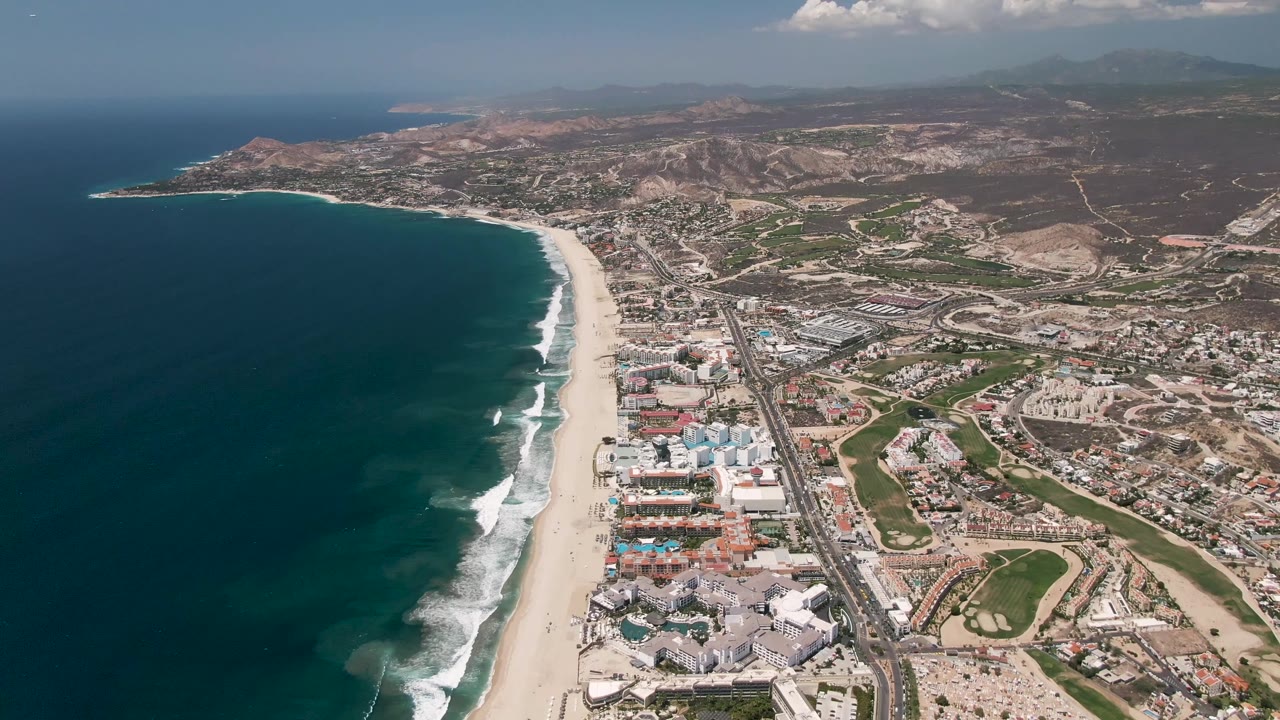 Aerial view over San Jose del Cabo with more open ocean visible at left, bright wave breaks along the beach, clusters of pale buildings, highway traffic, brown hillside terrain, and no visible people.