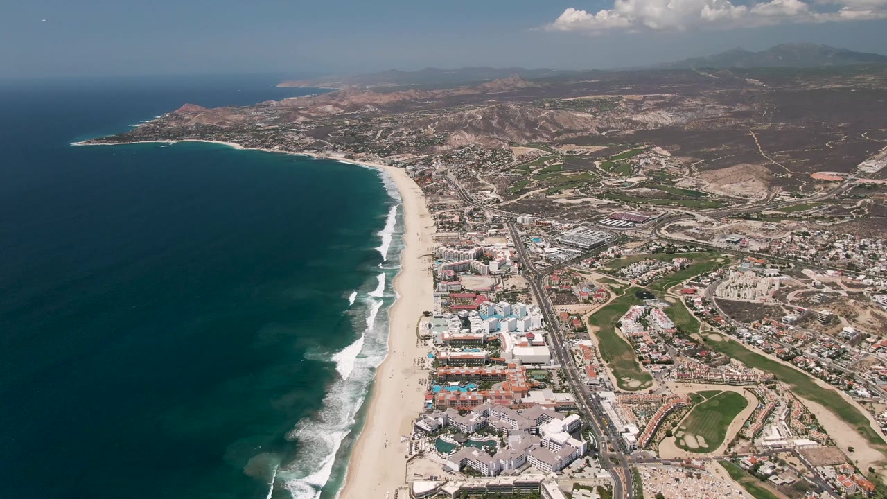 Aerial view of coastline with waves, beach, urban blocks, road, and hills.