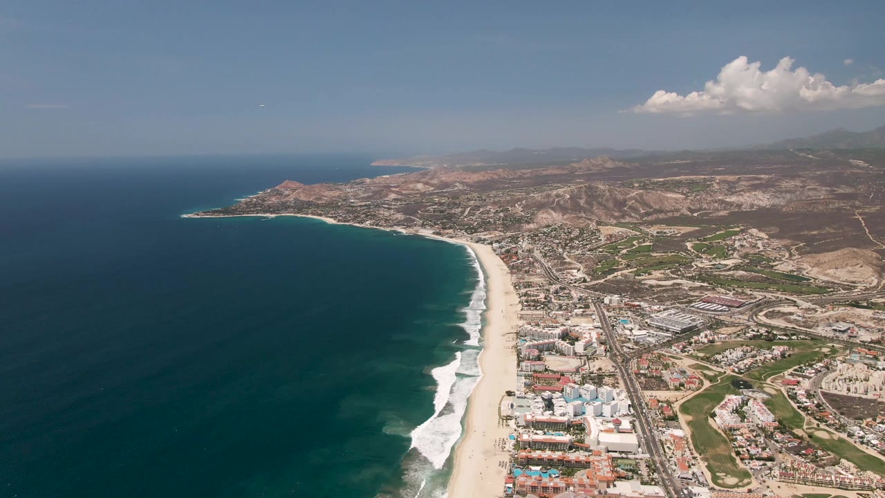 Aerial view of San Jose Del Cabo with deep blue ocean, a long sandy beach, coastal buildings, a road, dry hills, clouds, a small sky object, and no visible people.