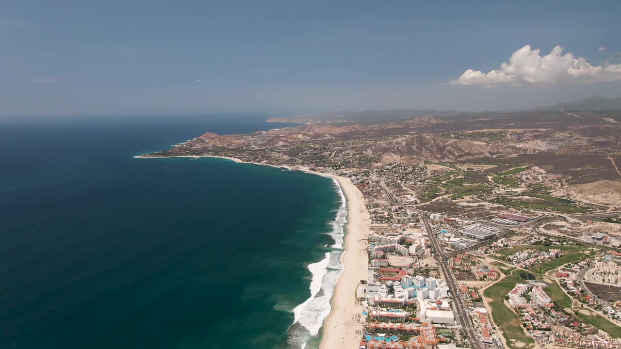 San Jose Del Cabo coastline from above, with the ocean occupying the left frame, white surf on the beach, developed shoreline, a road corridor, hills, clouds, and no visible people.