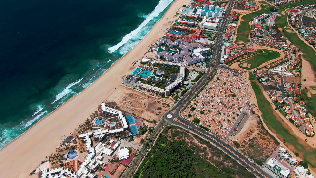 Aerial view of ocean waves, sandy beach, coastal road, buildings, pools, and golf greens.