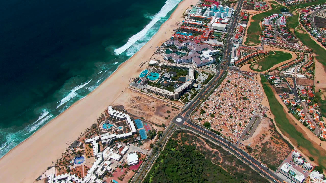 Second aerial frame in San Jose Del Cabo, Mexico, showing wave lines advancing toward the beach, resort blocks and turquoise pools near the road, golf-course grass to the inland side, and no visible people.