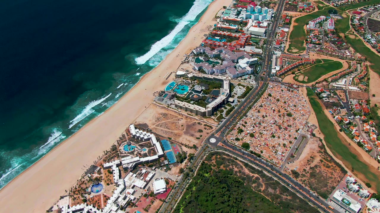 Third frame from the San Jose Del Cabo aerial shows darker ocean water, white wave crests near the sand, a central road, clustered resort buildings with blue pools, inland golf greens, and no visible people.