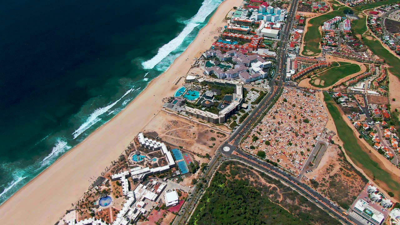 Mid-clip aerial frame over San Jose Del Cabo, Mexico, with the ocean and surf shifted along the left edge, empty sand below, road and resort structures in the middle, green areas inland, and no visible people.