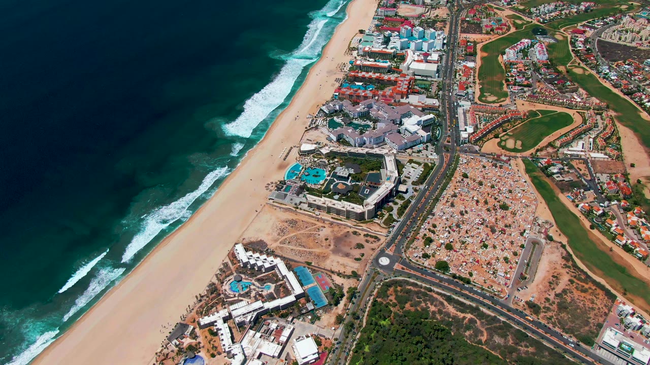 Drone-style view of San Jose Del Cabo with breaking waves on the left, a broad sand strip, a parallel road, clustered buildings, and green areas beyond the shoreline, with no visible people.
