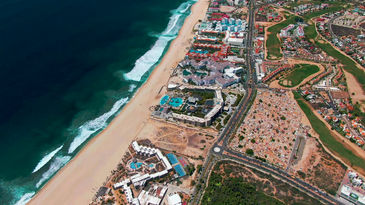 Coastal aerial frame with surf, beach, resort buildings, road, and green areas.
