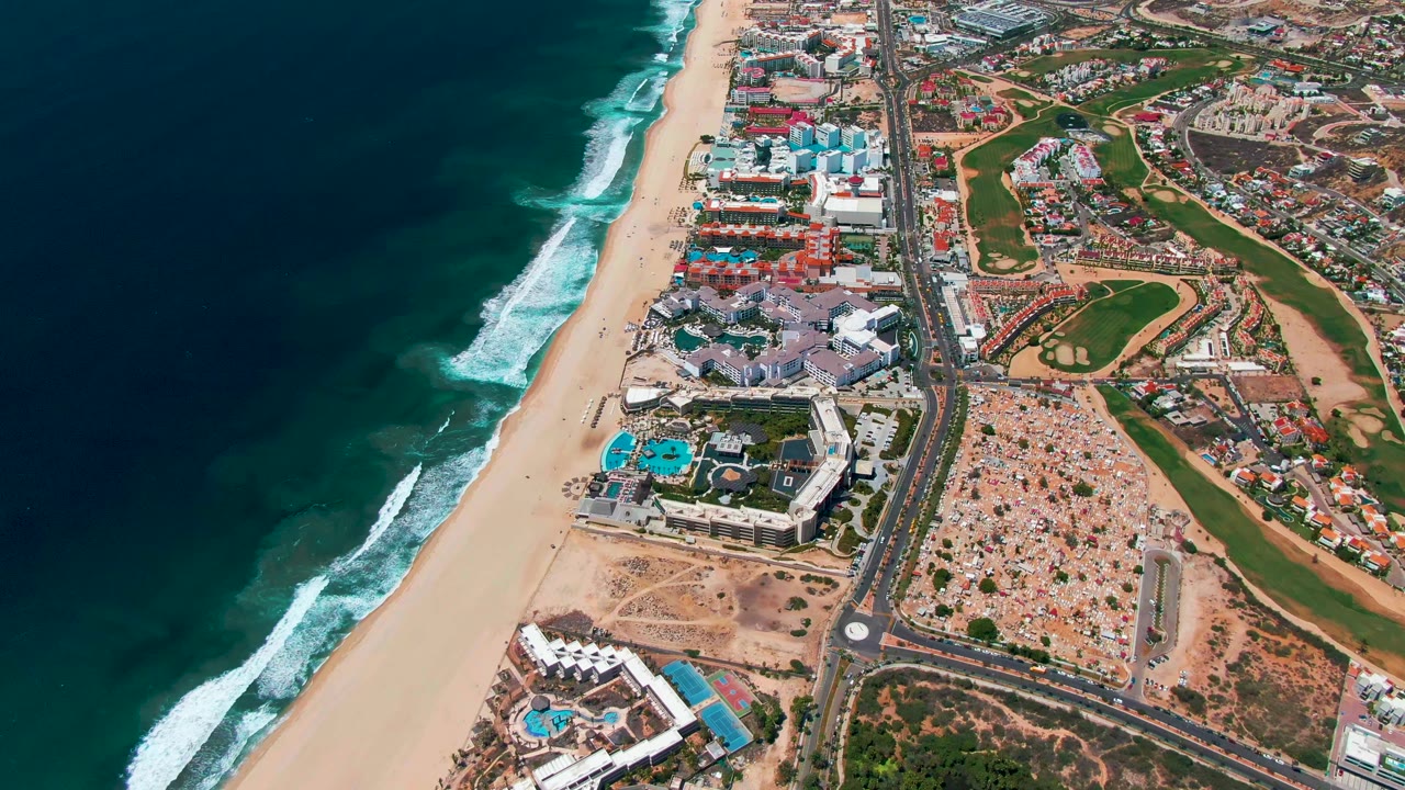 High aerial view over San Jose Del Cabo showing dark blue ocean, white surf, a sandy beach, resort pools, a parallel road, and inland golf course areas with no visible people.