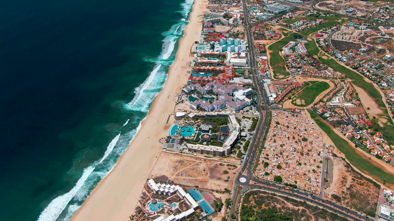Drone view of beach surf beside resort buildings and green golf fairways.